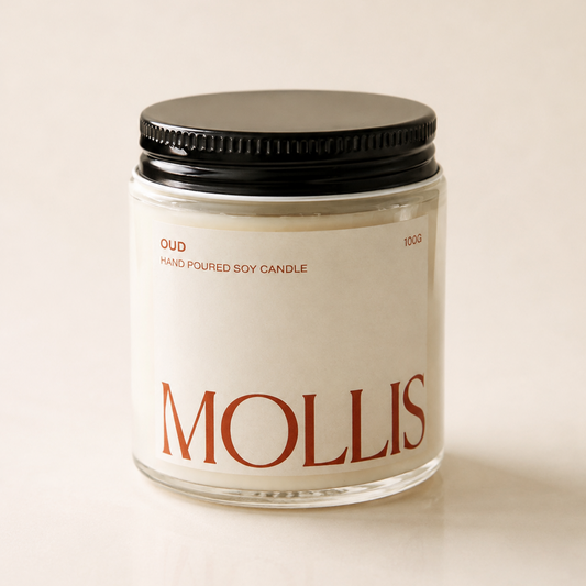 A glass jar candle with a black metal lid and a cream-colored label featuring the brand name 'MOLLIS' in red. The label also reads 'OUD' and 'HAND Poured SOY CANDLE'.