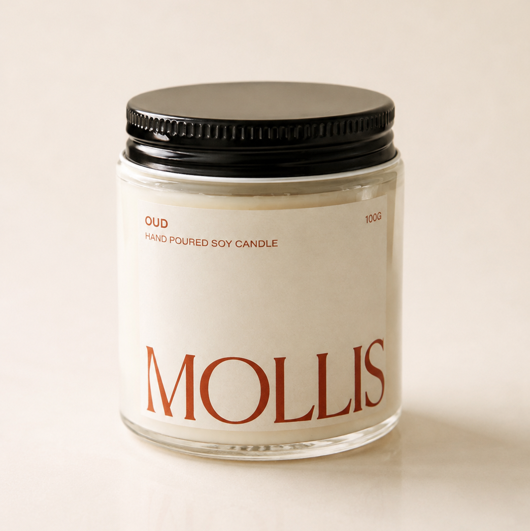 A glass jar candle with a black metal lid and a cream-colored label featuring the brand name 'MOLLIS' in red. The label also reads 'OUD' and 'HAND Poured SOY CANDLE'.